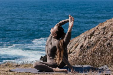 Dişi yogi, okyanus kenarındaki bir mindere uzanıyor. Etrafı kayalık arazi ve dalgalarla çevrili. Meditasyon ve rahatlama için huzurlu bir atmosfer yaratıyor.