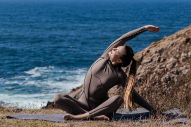 Dişi yogi, okyanus kıyısındaki bir mindere esneme hareketi yapıyor. Etrafı kayalık arazi ve dalgalarla çevrili. Huzurlu ve rahatlatıcı bir atmosfer yaratıyor.