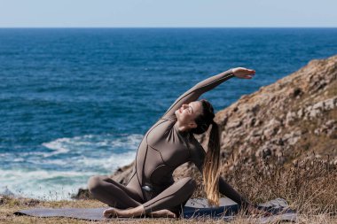 Dişi yogi, okyanusun kenarındaki bir mindere geriliyor. Arka planda kayalık bir manzara ve dalgalar var. Huzurlu ve meditasyon yapan bir atmosfer yaratıyor.