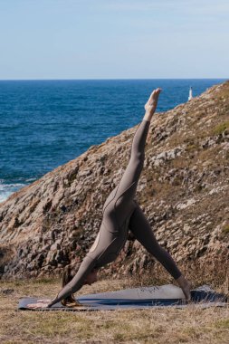 Yoga yapan bir kadın okyanusun kenarındaki bir paspasın üzerinde, kayalık bir çevrede, huzurlu bir atmosferde, rahatlama ve refahı destekleyen, aşağı bakan bir köpek pozu veriyor.