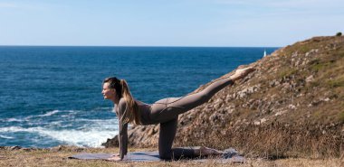 Dişi yogi, doğal güzelliklerle çevrili, okyanus kıyısındaki bir mindere gerilme pozu verir. Huzurlu bir ortamda rahatlama ve zihinsel berraklığı teşvik eder.