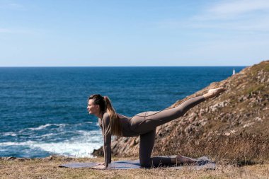 Dişi yogi, dalgaların ve uçurumların manzaralı zeminiyle okyanus kıyısındaki bir mindere esneme pozu veriyor. Rahatlamayı ve iç huzuru teşvik ediyor.