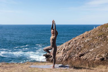 Dişi yogi açık havada dengeleyici bir duruş sergiliyor, arka planda okyanus dalgaları ve kayalık bir manzara, barışçıl ve uyumlu bir atmosfer yaratıyor.