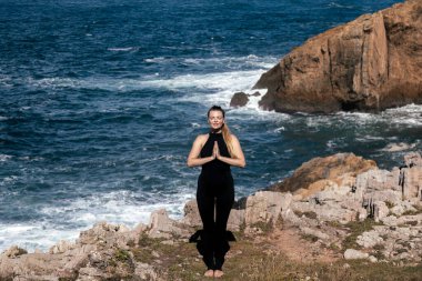Meditasyon yapan ve esneme hareketleri yapan kadın figürü kayalık kıyılarda, okyanus dalgaları yakınlarda çarpışırken, sakin ve huzurlu bir atmosfer yaratıyor.