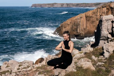 Meditasyon yapan ve esneme hareketleri yapan kadın figürü kayalık kıyılarda, okyanus dalgaları yakınlarda çarpışırken, sakin ve huzurlu bir atmosfer yaratıyor.