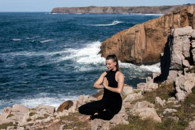 Meditasyon yapan ve deniz kenarındaki engebeli kayalara gerinen kadın figürü, çarpan dalgalar ve uçurumlar farkındalık için sakin bir atmosfer yaratıyor.