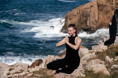 Dişi meditasyon ustası, deniz kıyısındaki kayaların üzerinde bağdaş kurarak oturur, elleri dua pozisyonunda, dalgalar arka planda çarpar ve kayalıklar barışçıl bir atmosfer yaratır.