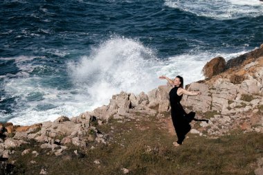 Kadın figürü meditasyonla meşgul ve kayalık kıyılarda geriniyor, çarpan dalgalar ve doğal güzelliklerle çevrili, rahatlamayı ve farkındalığı teşvik ediyor.
