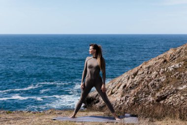 Kadın yoga uygulayıcısı, doğal bir ortamda, okyanus dalgaları ve kayalık araziyle çevrili bir mindere güvenerek duruyor, rahatlamayı ve odaklanmayı teşvik ediyor.