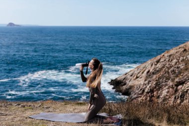 Kadın pratisyen, meditasyon yapar ve doğal güzelliklerle çevrili okyanus kenarında bir yoga minderine uzanır. Rahatlamayı ve huzuru teşvik eder.
