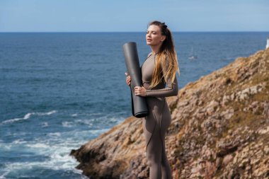 Kadın yoga uygulayıcısı kayalık sahilde duruyor, arka planda okyanus dalgaları olan minderi tutuyor, vücut ve doğa arasındaki uyumu özenli esneme yoluyla gösteriyor.