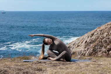 Dişi yogi, okyanusun kenarındaki bir mindere uzanıyor, doğayı ve sükuneti kucaklıyor, arka planda dalgalar çarpıyor, barışçıl atmosferi güçlendiriyor.