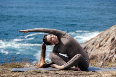 Dişi yogi, okyanusun kenarındaki bir mindere uzanıyor. Dalgalar ve kayalık arazi onu çevrelemiş. Meditasyon için sakin ve huzurlu bir atmosfer yaratıyor.