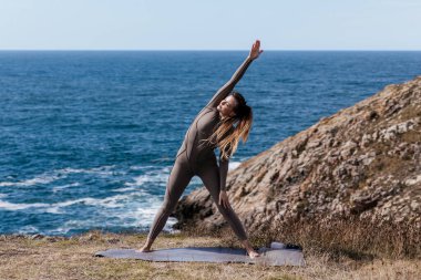 Kadın yoga uygulayıcısı okyanus kıyısında bir mindere geriliyor. Arka planda dalgalar ve kayalık arazi barışçıl ve doğal bir atmosfer yaratıyor.