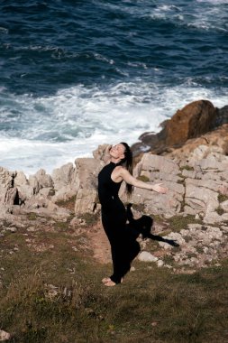 Kadın yoga uygulayıcısı kayalık sahilde duruyor, kolları tamamen açık, doğayla iç içe, okyanus dalgaları ve kayalık arazi ile çevrili, barış ve sükuneti somutlaştırıyor.