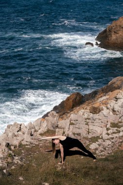 Kadın yoga uygulayıcısı okyanus kıyısındaki kayalık arazide dalgaların sahile vurmasıyla sakin ve huzurlu bir atmosfer yaratıyor.