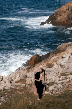 Dişi yogi, okyanus dalgaları ve doğal manzarayla çevrili kayalık kıyılarda esniyor. Huzurlu bir ortamda rahatlama ve farkındalığı teşvik ediyor.
