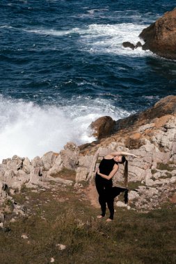 Dişi yogi kayalık arazide esneme hareketi yapıyor. Yakınlarda okyanus dalgaları çarpıyor. Huzurlu bir farkındalık ve rahatlama atmosferi yaratıyor.