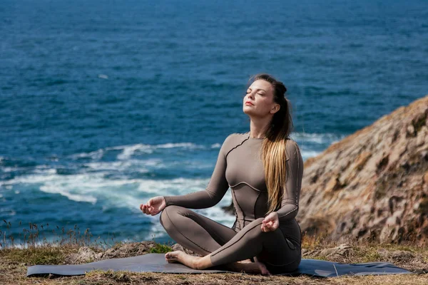 Dişi yogi okyanus kıyısında bir minder üzerinde meditasyon yapıyor. Kayalık arazi ve arka planda hafif dalgalar var. Huzurlu ve rahatlatıcı bir atmosfer yaratıyor.