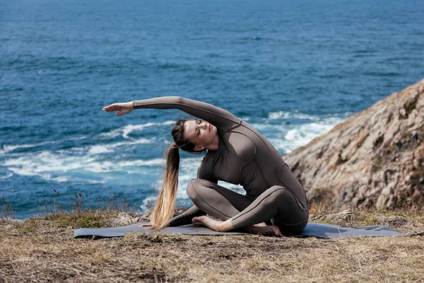 Dişi yogi, okyanus kıyısındaki bir mindere uzanıyor, esneklik ve dengeyi doğal bir ortamda gösteriyor, rahatlık ve rahatlığı teşvik ediyor.