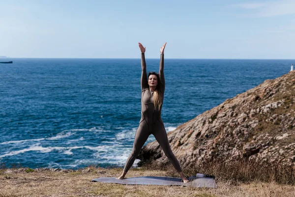 Dişi yogi, okyanus kıyısındaki bir mindere esneme pozu veriyor. Arka planda kayalık arazi ve sakin su var. Rahatlamayı ve refahı teşvik ediyor.