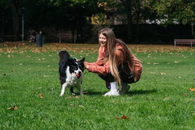 Uzun saçlı bir kadın, sonbahar yapraklarıyla çevrili canlı yeşil bir parkta, siyah ve beyaz bir köpekle şakalaşarak bir mutluluk anı yakalıyor.