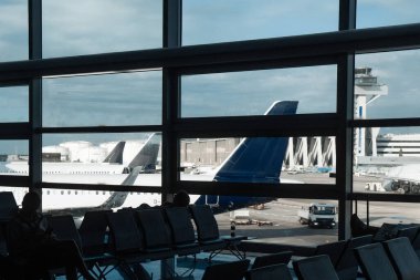 Havaalanı terminalinin içinde, pistte park halindeki uçakları gösteren geniş pencereler ve bekleme alanında oturan yolcular, seyahat atmosferini çağrıştırıyor.