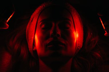 Kozmetik bilimci yüz tazeleme için LED ışık terapisi uyguluyor, parlak kırmızı ışıklar ve rahatlatıcı bir atmosfer vurguluyor, cilt sağlığı ve güzelliği artırıyor