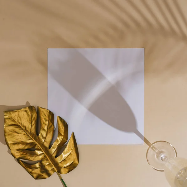 金色のモンテラの葉と砂のベージュの背景にヤシの影を持つ白いペーパーカードとシャンパングラスの最小限のトップビュー 夏のクリエイティブコンセプト — ストック写真