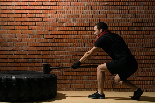 man with a hammer in his hand beats on a tire, training crossfit against a brick wall