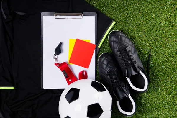 soccer ball, boots, a recording tablet, a whistle and two penalty cards for a judge, against the background of a sports T-shirt and grass
