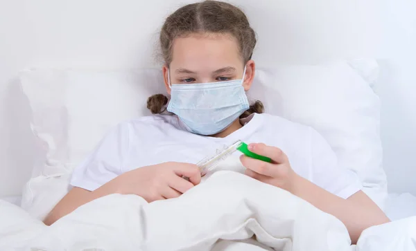 一个在床上的女孩 在一个医学面具检查身体的温度 拿着温度计 — 图库照片