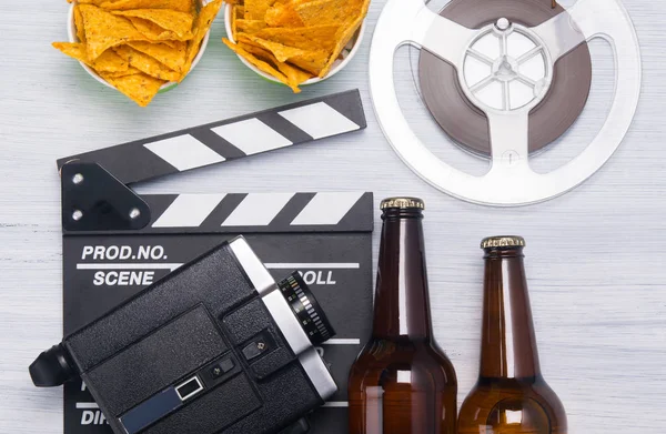 two bottles of beer, a bucket of nachos, a movie clapper with an old video camera and a film strip, on a gray background