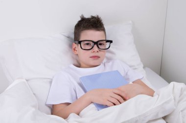 Gözlüklü erkek yatakta yatıyor ve bir kitap elinde tutar