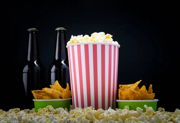 two glasses of popcorn and nachos, on a black background, with two bottles of beer