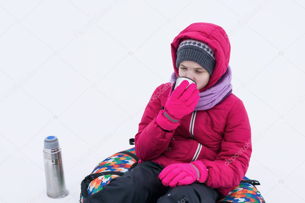 girl in a red jacket sits in the snow and drinks warm tea from a thermos