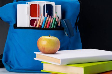 mavi bir sırt çantası içinde renkli kalemler ve boyalar ve kitap ve snack-elma vardır
