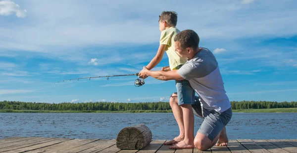 桟橋の父親は 青い湖と空に対して 釣りのために釣り竿を維持するために彼の息子を助けます — ストック写真
