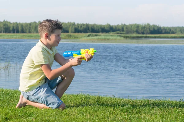 少年は芝生の上に座っているし 夏の湖のピストルからの水を撮影 — ストック写真