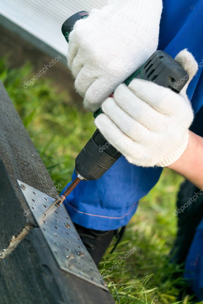 a man in white gloves spins a screw into a black wooden beam with an electric screwdriver
