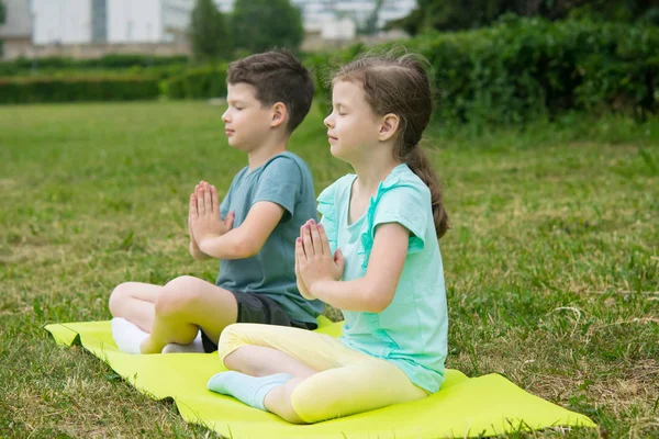 boy and girl do yoga, eyes closed, outdoors, on a background of green grass, sitting on a gymnastic mat, side view