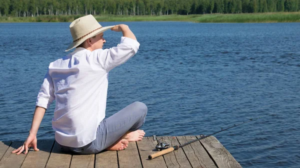 埠頭に座って 遠くを見て 湖に逆らって釣り竿の横に横たわっている男 — ストック写真