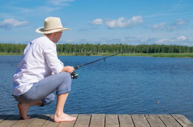 bir şapkalı bir adam, iskelede oturan, bir olta tutan ve balıkçılık, mavi gökyüzü ve göle karşı