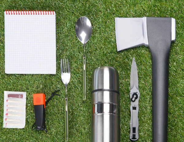 on the green lawn, neatly laid out items for wild outdoor recreation, matches, ax, thermos, fork, spoon, flashlight, knife and a clean Notepad for writing