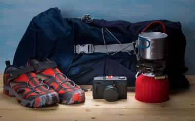 ahşap masa ve mavi arka plan, gezgin seti, büyük mavi sırt çantası, rahat ayakkabı, kamera ve gaz brülör