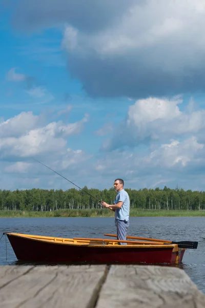 黄色い眼鏡をかけた男 手漕ぎボート 釣り竿を持ち 桟橋と美しい景色の背景に魚を捕まえる — ストック写真