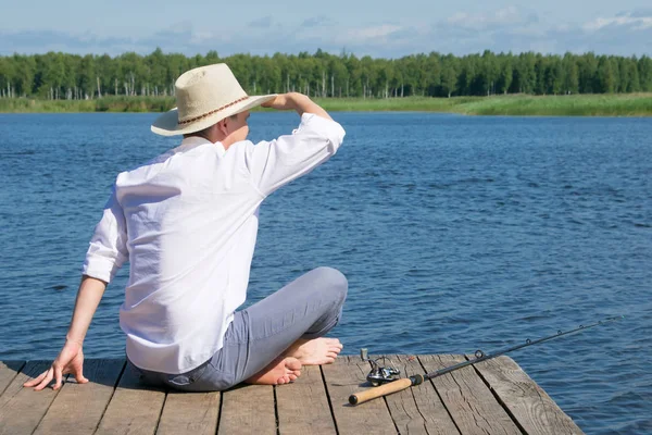 帽子をかぶった男が桟橋に座り 遠くに座り 釣り竿の横に横たわり 青い空と湖に向かう — ストック写真