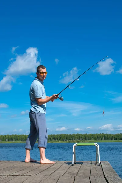 桟橋に黄色い眼鏡をかけた男は 青い空と湖に対して 釣りのために釣り竿を投げる — ストック写真