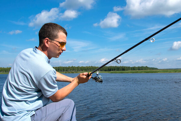 a man in yellow glasses on the pier, throws spinning, fishing, against the blue lake and sky