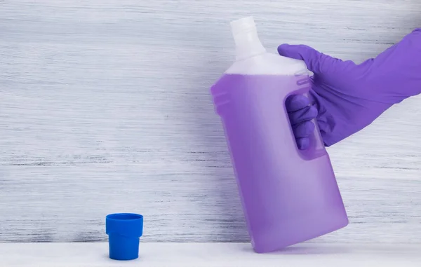 hand in a purple, protective glove holding an open bottle of cleaning fluid, on a gray background, with space for inscription